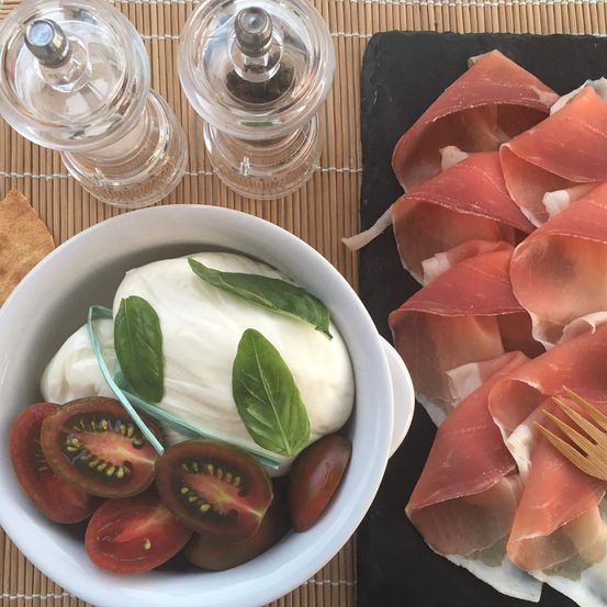 Ein weißer Bowl mit Tomaten, Basilikum und Mozzarella liegt neben einer schwarzen Platte mit Prosciutto. Zwei Glas-Shaker sind auf dem Tisch.