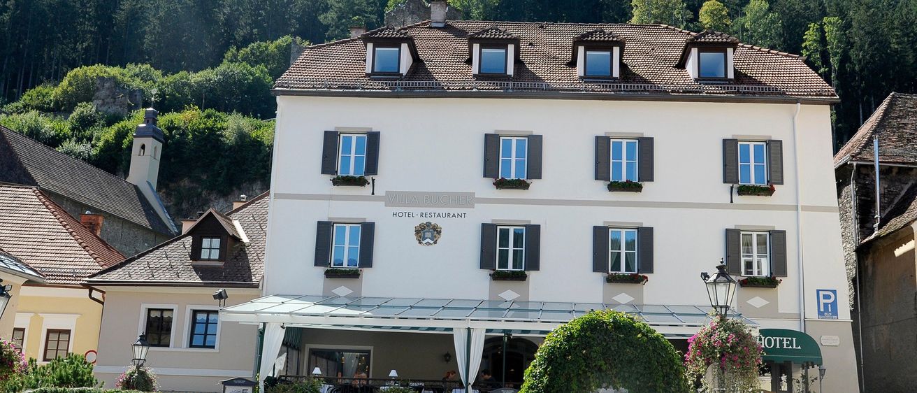 Ein weißes Gebäude mit braunem Dach und mehreren Fenstern. Das Gebäude heißt Hotel-Restaurant. Das Gebäude hat einen Eingang mit Stufen und einem grünen Bogen.
