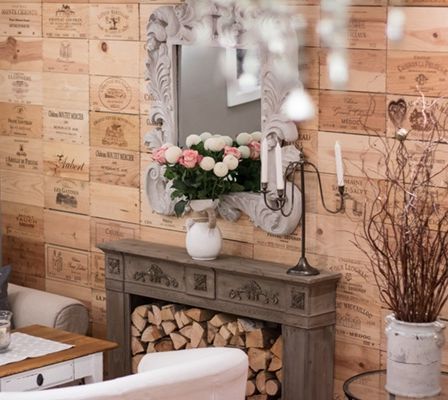 Ein gemütlicher Raum mit Holzwänden, die mit Weinhekten verziert sind. Ein Kamin mit Brennholz und eine Vase mit Blumen. Ein Spiegel mit einem verzierten Rahmen reflektiert eine weiße Kerze.
