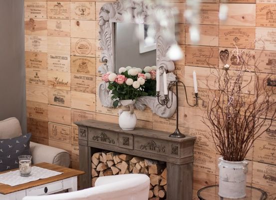 Ein gemütlicher Raum mit Holzwänden, die mit Weinhekten verziert sind. Ein Kamin mit Brennholz und eine Vase mit Blumen. Ein Spiegel mit einem verzierten Rahmen reflektiert eine weiße Kerze.