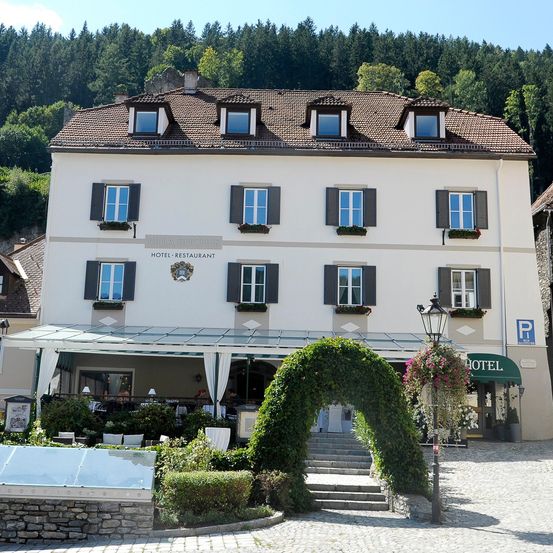 Ein weißes Hotel mit einem braunen Dach und vielen Fenstern, umgeben von Grün und Bergen.