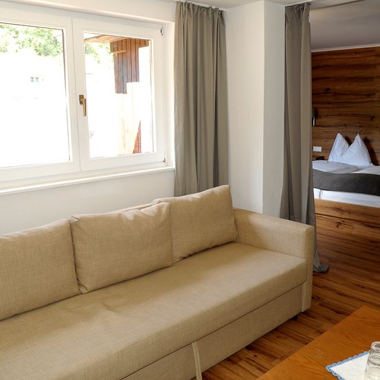 Innenansicht eines Hotelzimmers mit einem beigen Sofa, das zu einem großen Fenster und einer Holzwand ausgerichtet ist. Hinter dem Sofa befindet sich ein Holzbett mit zwei Kissen.