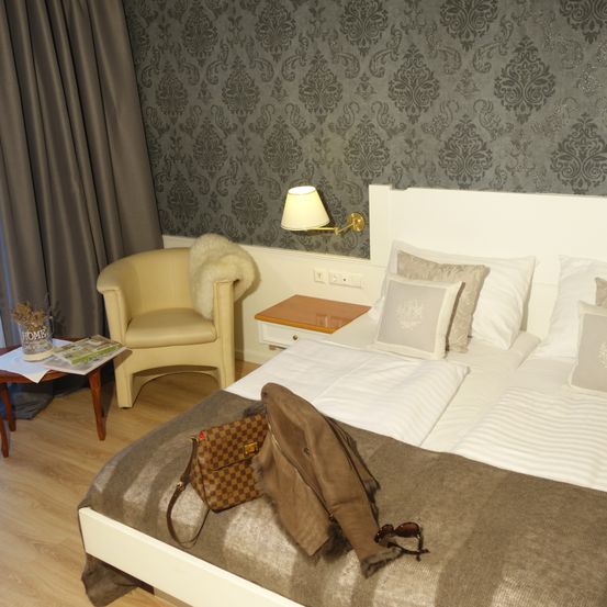 Ein Zimmer mit einem Bett, einem Stuhl und einem Nachttisch. Das Bett hat ein weißes Kopfteil und mehrere Kissen. Eine Jacke, eine Handtasche und eine Brille liegen auf dem Bett. Der Stuhl hat ein Fellkissen.