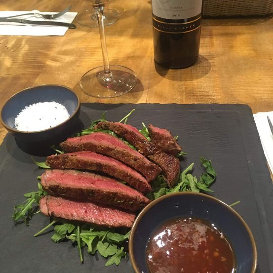Ein Gericht mit Steak, Rucola, einer Schüssel Sauce und einer Flasche Wein auf einem Holztisch mit einem schwarzen Schneidebrett.