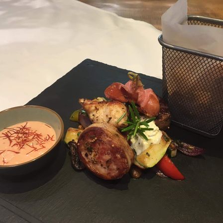 Ein Gourmetgericht mit einem Stück Fleisch, Gemüse und einer Beilage Dip. Ein Korb aus Edelstahl mit einer Serviette daneben.