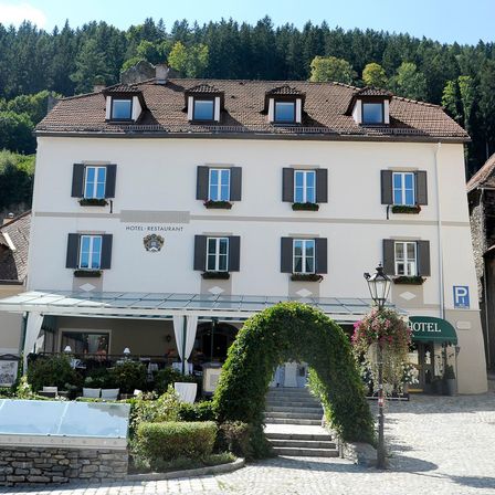 Ein weißes Hotel mit einem braunen Dach steht in einem Gebirgsgebiet. Es hat mehrere Fenster mit schwarzen Fensterläden. Der Eingang ist durch ein Vordach und einen Bogen gekennzeichnet.