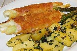 Ein weißer Teller enthält gebratenen Spargel, der in Schinken eingewickelt ist, begleitet von einer Schale Kartoffelsalat mit Kräutern.