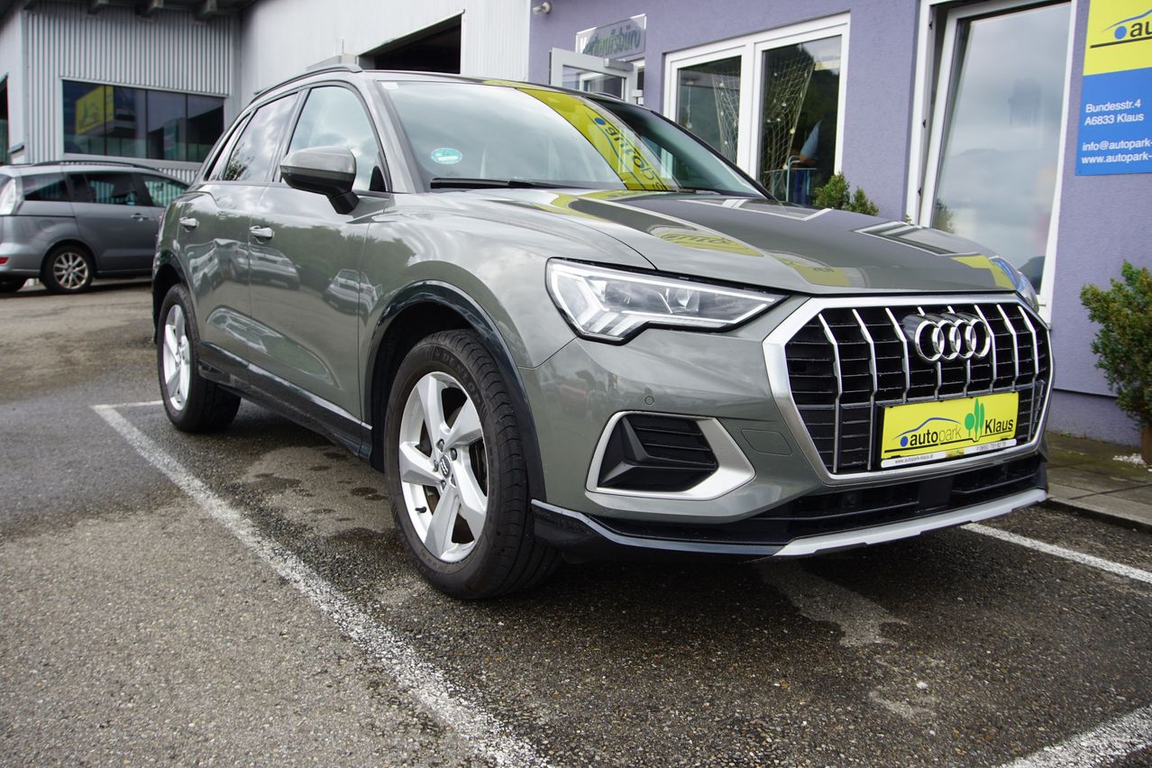 Ein grauer Audi SUV ist vor einem Gebäude mit Glasfenstern und einem gelben Nummernschild geparkt.