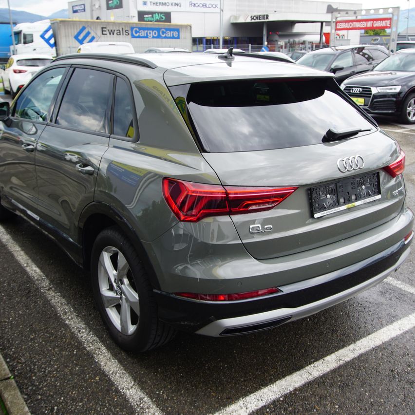 Die Heckansicht eines grauen Audi Q3 steht auf einem Parkplatz neben anderen Autos. Das Auto hat ein Nummernschild und einen schwarzen Spoiler.