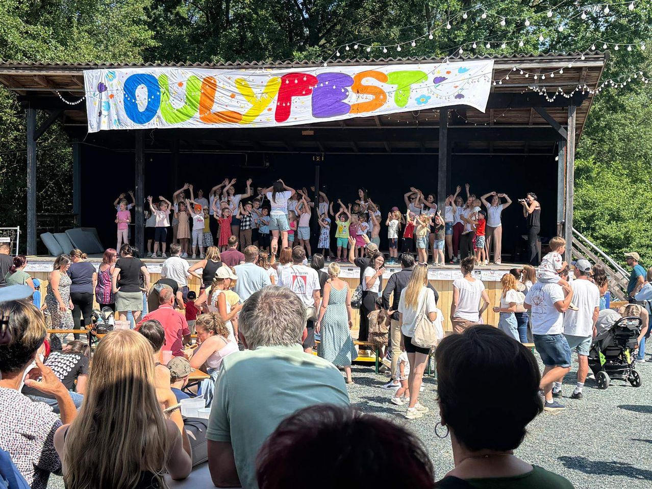 Eine Menschenmenge, bestehend aus Kindern und Erwachsenen, versammelt sich bei einem Open-Air-Festival. Auf der Bühne stehen eine Gruppe von Kindern und Erwachsenen mit erhobenen Händen. Ein Banner mit der Aufschrift 'OULYPFEST' hängt darüber.