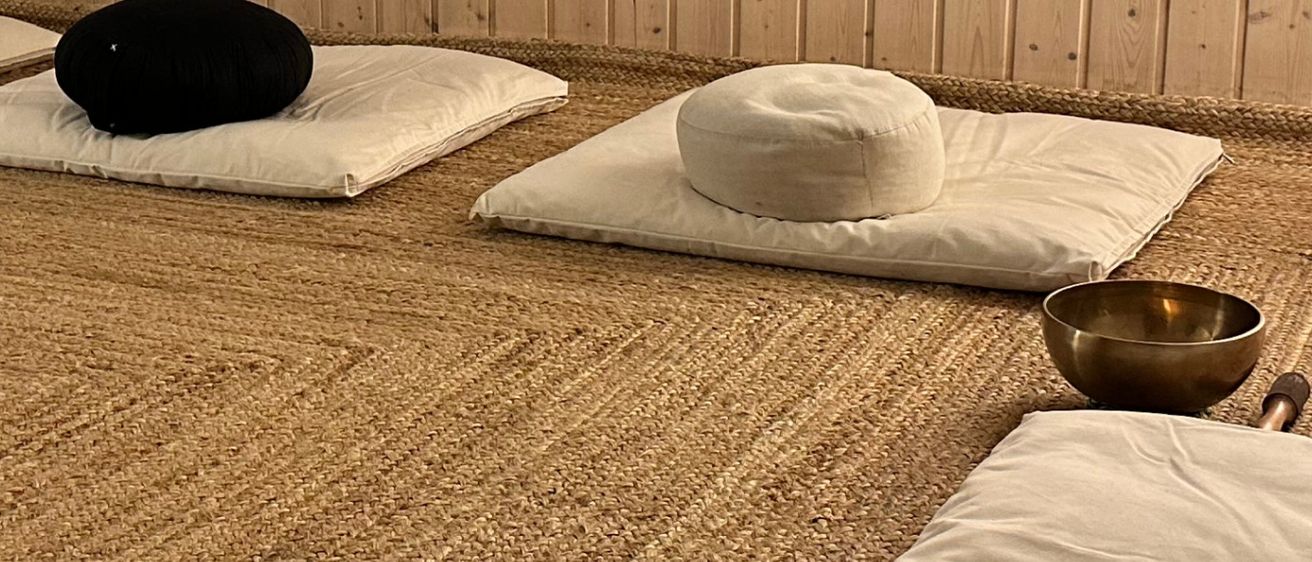 Meditationsraum mit vier gepolsterten Sitzen, Schalen, einer Lampe und einem kleinen Tisch mit einer roten Lampe.