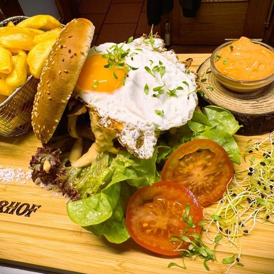 Ein Gourmet-Burger mit Spiegelei, Salat, Tomaten und Sprossen auf einem Holzbrett. Pommes und eine Schüssel Sauce sind auf der Seite.
