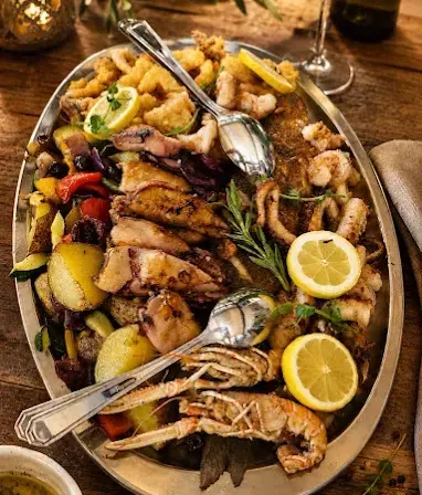 Ein großer Teller mit Meeresfrüchten und Gemüse, darunter Garnelen, Jakobsmuscheln und Zitronenscheiben, garniert mit Kräutern und serviert mit einer Beilage zum Dippen.