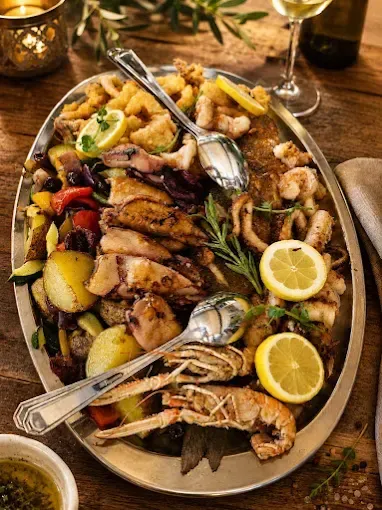 Ein großer Teller mit Meeresfrüchten und Gemüse, darunter Garnelen, Jakobsmuscheln und Zitronenscheiben, garniert mit Kräutern und serviert mit einer Beilage zum Dippen.