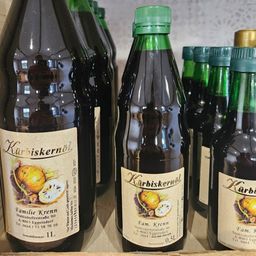 Mehrere Flaschen Kürbiskernöl sind auf einem Holzregal angeordnet, jede mit einem grünen Deckel und Etikett mit einer Illustration eines Kürbisses.