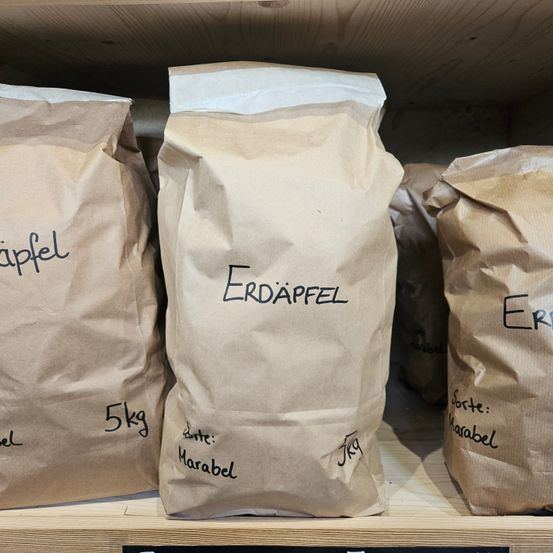 Drei braune Papiertüten, die auf einem Holzregal gestapelt sind. Die mittlere Tüte ist mit 'ERDAPFFEL' und 'Marabel' beschriftet. Die linke Tüte ist mit '5kg' und die rechte Tüte mit 'Marabel' beschriftet.