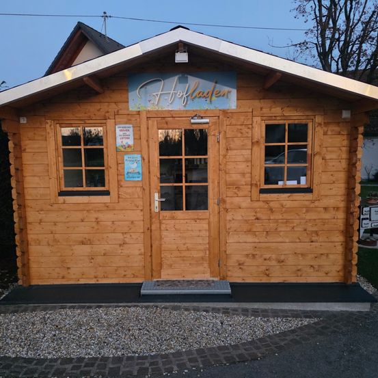 Ein Holzgebäude mit einem Schild oben, das 'Hoflader' besagt, drei Fenster und eine Tür. Das Gebäude befindet sich auf einer Kiesauffahrt, und es gibt ein Schild mit Text auf der linken Seite.