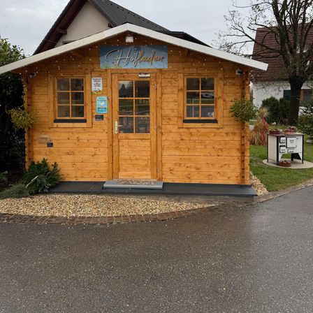 Ein kleines Holzhaus mit einem blauen Banner mit der Aufschrift Hoffelder, gelegen an einer Straße mit einem Rasenbereich und einem Baum.