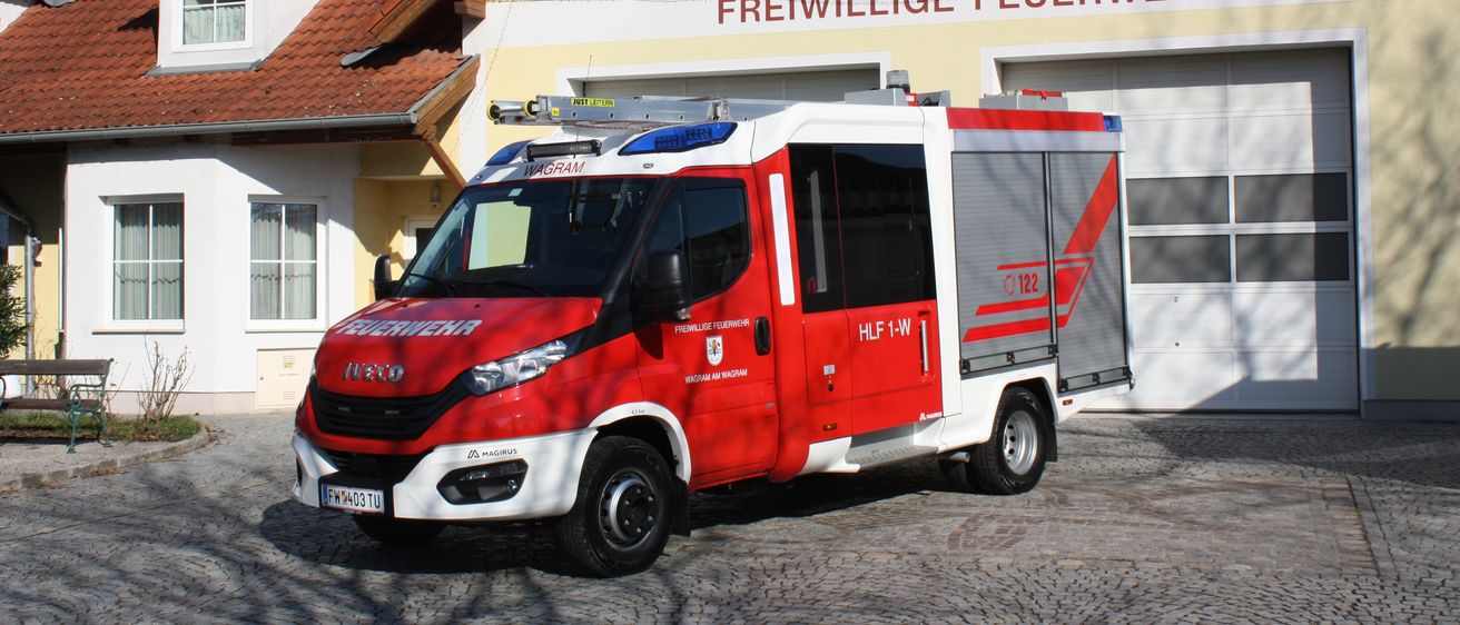 Ein roter und weißer Feuerwehrwagen steht vor einem Gebäude mit der Aufschrift 'Freiwillige Feuerwehr'.