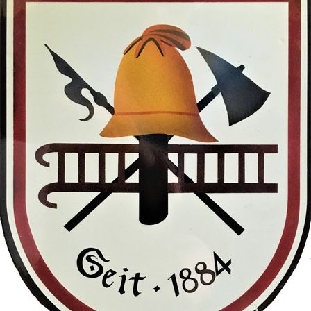 Das Emblem zeigt einen Feuerwehrhelm, eine Axt und eine Leiter. Das Emblem hat die Form eines Schildes mit dem Text 'Seit 1884' unten.