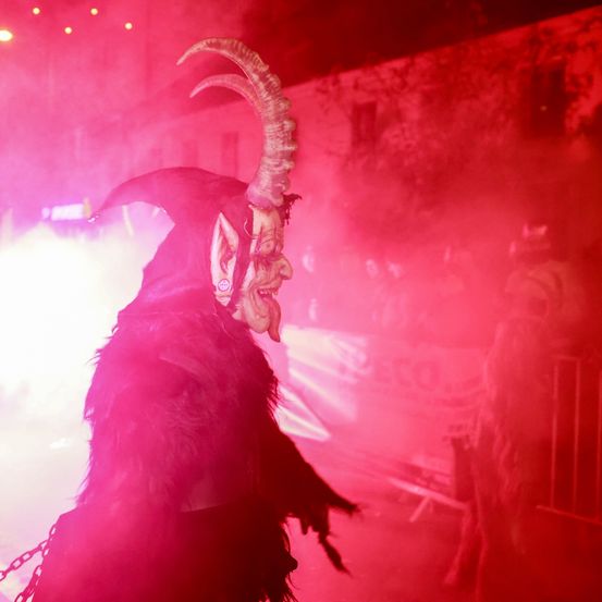Eine Person in einer Hornmaske steht vor einer rosa beleuchteten Wand, trägt Ketten und ein schwarzes Kostüm. Im Hintergrund sind verschwommene Figuren und roter Rauch zu sehen.