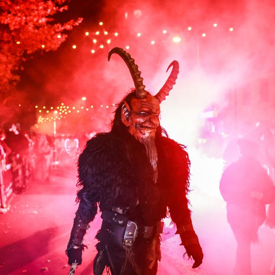 Eine Person in Teufelskostüm mit Hörnern und roter Maske geht nachts durch eine Straße. Eine Menge beobachtet, und Feuerwerke erhellen den Hintergrund.