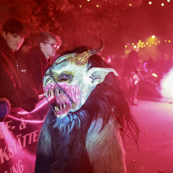 Eine Person mit einer hörnertragenden, gruseligen Maske steht in der Nacht vor einer Menge. Die Person ist in einem langen pelzigen Kostüm gekleidet. Der Hintergrund ist dunkel mit roten Lichtern.