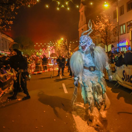 Eine Person in einem Dämonenkostüm geht bei Nacht die Straße entlang, umgeben von Menschen und Gebäuden mit beleuchteten Schildern.