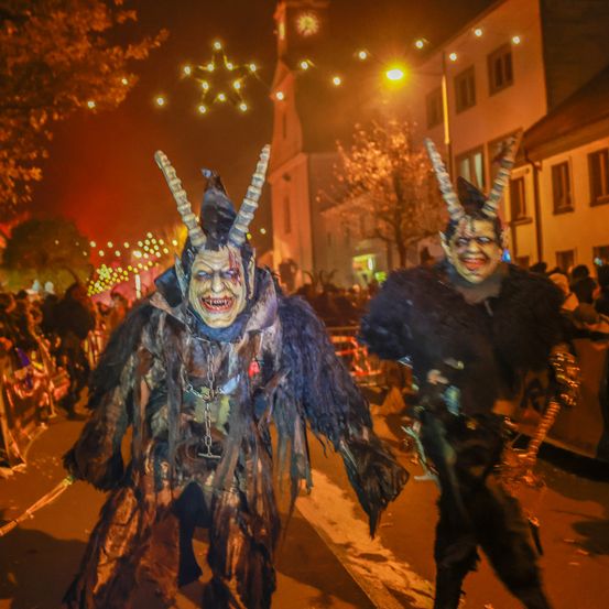 Zwei Personen in Hörnerkostümen gehen während eines nächtlichen Festivals durch eine Straße. Sie sind von einer Menge umgeben, mit Feuerwerk und Gebäuden im Hintergrund.