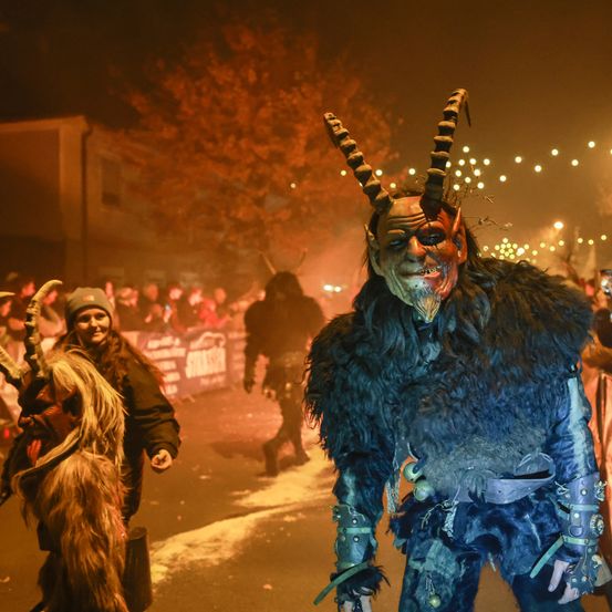 Ein nächtliches Festival mit Menschen in aufwendigen Kostümen, einer trägt eine gehörnte Maske, während andere im Hintergrund von roten Lichtern und Rauch beleuchtet sind.