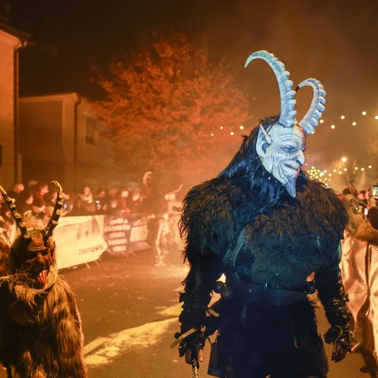 Eine Person in einer Ziegenmaske geht bei Nacht eine Straße entlang, umgeben von einer Menge. Im Hintergrund befindet sich ein Baum mit leuchtenden Lichtern.
