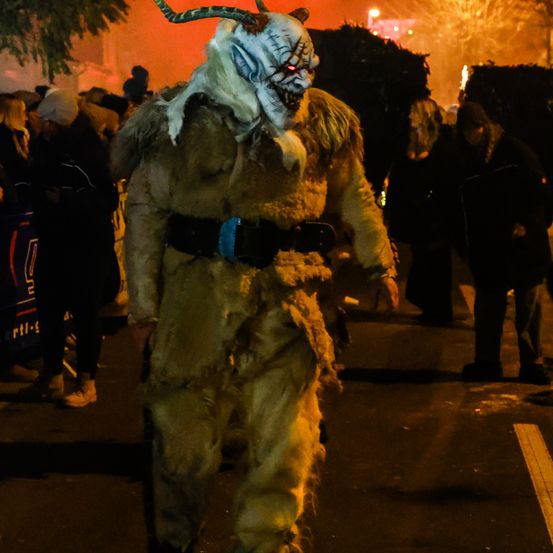 Eine Person in einem Dämonenkostüm geht bei Nacht durch eine Menge, mit roter Beleuchtung und Bäumen im Hintergrund.