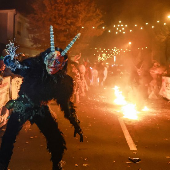 Eine Person in einem Teufelskostüm mit Hörnern und einer Maske geht auf einer Straße mit Feuer im Hintergrund.