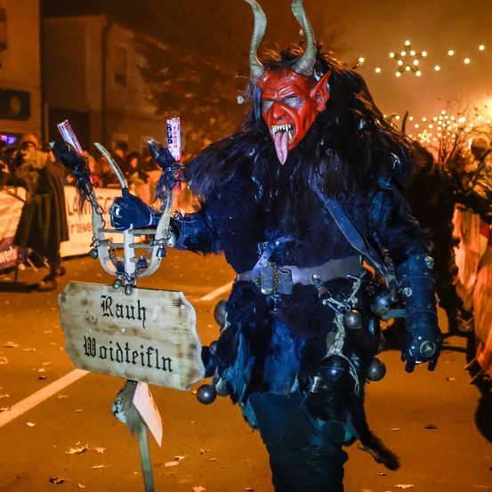 Eine Person als Teufel verkleidet bei einer Parade, hält einen Dreizack und ein Schild mit der Aufschrift 'Rauhn Woidtreifln', mit Menschen und Gebäuden im Hintergrund.