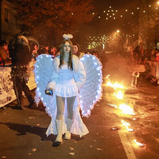 Eine Frau in einem Engelkostüm läuft in einer nächtlichen Parade, beleuchtet durch ihre leuchtenden Flügel. Sie steht auf der Straße, während die Leute zusehen, und im Hintergrund brennen Feuer.