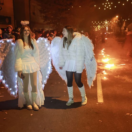 Zwei Frauen, die als Engel verkleidet sind, stehen bei Nacht auf einer Straße mit brennenden Feuern und einer Menschenmenge hinter ihnen.