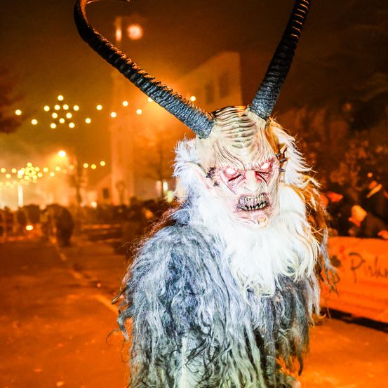 Eine Person in einer gehörnten Maske und einem rauen Kostüm steht nachts auf einer Straße, mit einer Menge und Lichtern im Hintergrund.