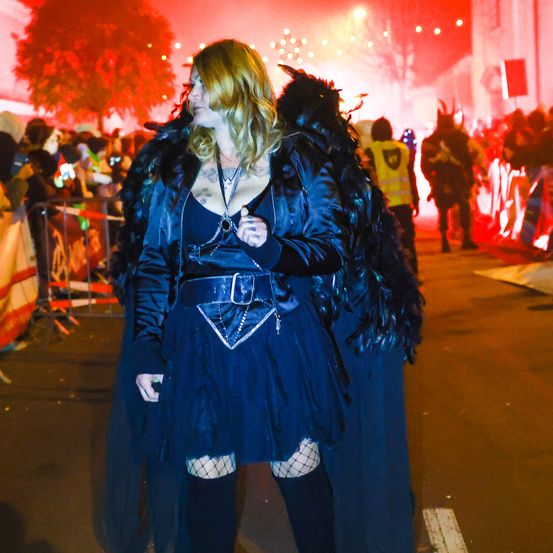 Eine Frau in einem blauen Kostüm mit Flügeln geht bei Nacht die Straße entlang, mit einer Menge hinter ihr.