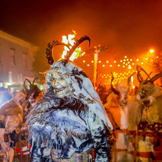 Ein nächtliches Festival mit einer Menschenmenge in Tierkostümen und Hörnern, die vor einem Lagerfeuer stehen. Der Hintergrund ist schwach beleuchtet, auch andere Menschen sind in Kostümen gekleidet.