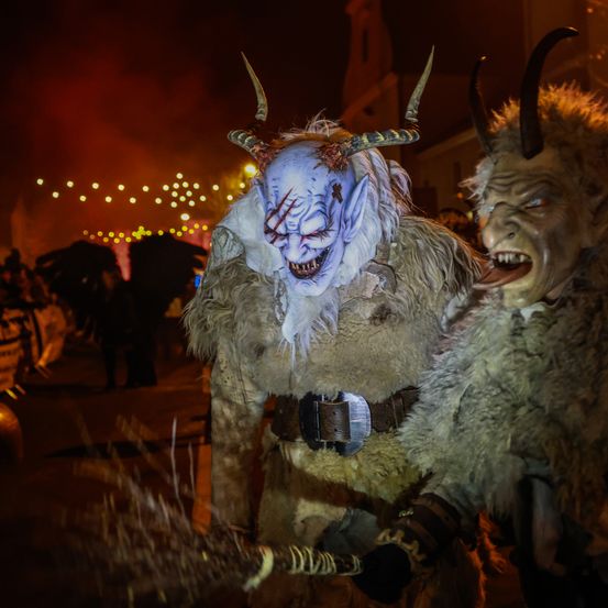 Zwei kostümierte Figuren, möglicherweise Kobolde, stehen bei Nacht zusammen. Sie tragen pelzige Kostüme, wobei eine Hörner und eine weiße Maske hat und die andere ein grünes Gesicht zeigt. Im Hintergrund sind verschwommene Menschen und Lichter zu sehen.