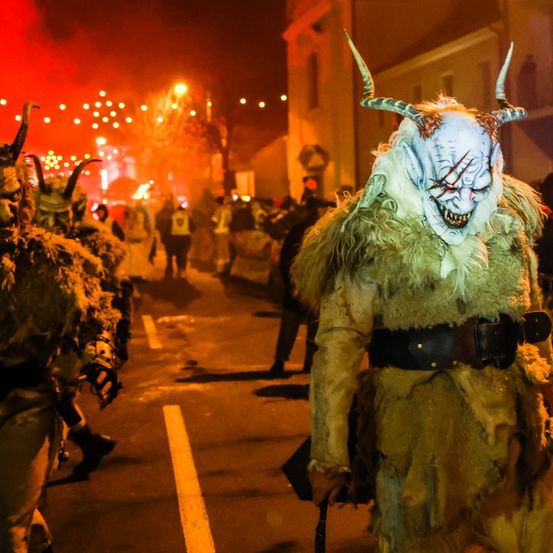 Eine Gruppe von Menschen in gruseligen Kostümen mit Hörnern geht nachts auf einer Straße. Die Straße ist mit roten Lichtern und Feuerwerk dekoriert.
