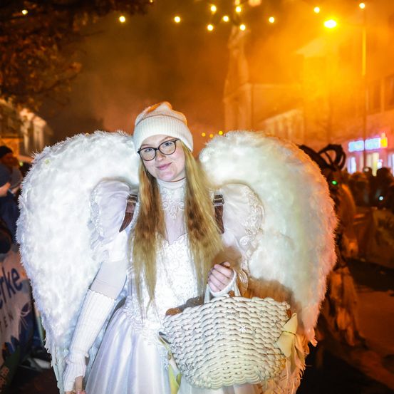 Eine Frau in einem Engelskostüm hält einen Korb auf einer nächtlichen Stadtstraße, umgeben von einer festlichen Atmosphäre und verschwommenen Figuren im Hintergrund.