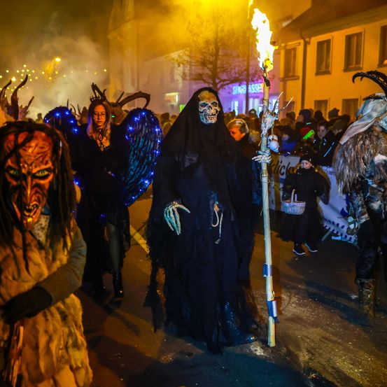 Menschen in Kostümen ziehen bei Nacht eine Straße entlang, halten eine Fackel und andere Requisiten. Sie nehmen an einem Halloween-Umzug teil.