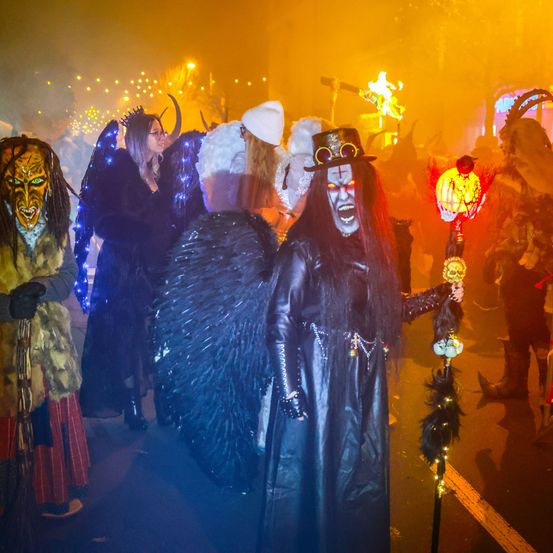 Ein Halloween-Umzug mit Menschen in Kostümen, einer hält eine leuchtende Laterne. Der Hintergrund ist rauchig mit Feuerwerk.