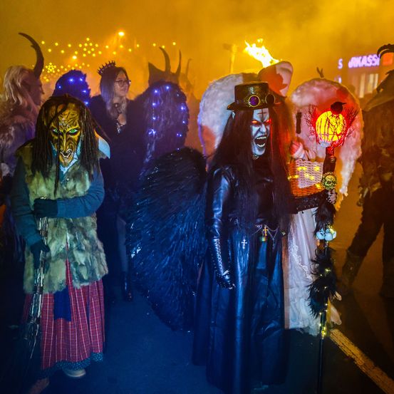 Eine Gruppe von Menschen in verschiedenen Kostümen, einschließlich Masken und Flügeln, zieht bei Nacht durch einen Umzug. Der Hintergrund ist mit gelben und orangefarbenen Lichtern verschwommen.