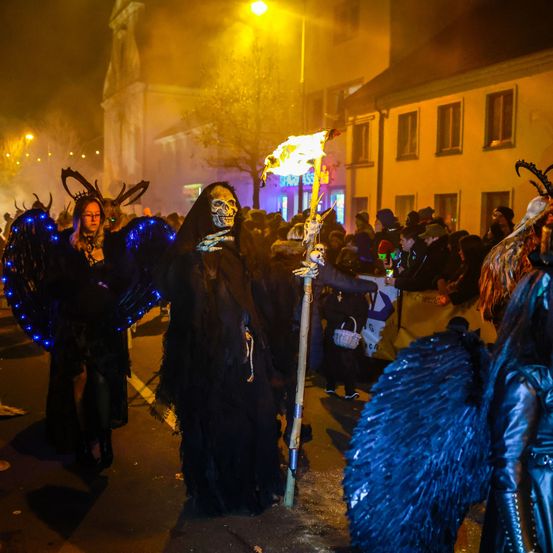 Bei Nacht marschieren eine Gruppe von Menschen in Kostümen auf der Straße, einer hält eine flammende Fackel. Die Kostüme umfassen Masken und Flügel. Die Straße ist dunkel mit Gebäuden und Bäumen im Hintergrund.