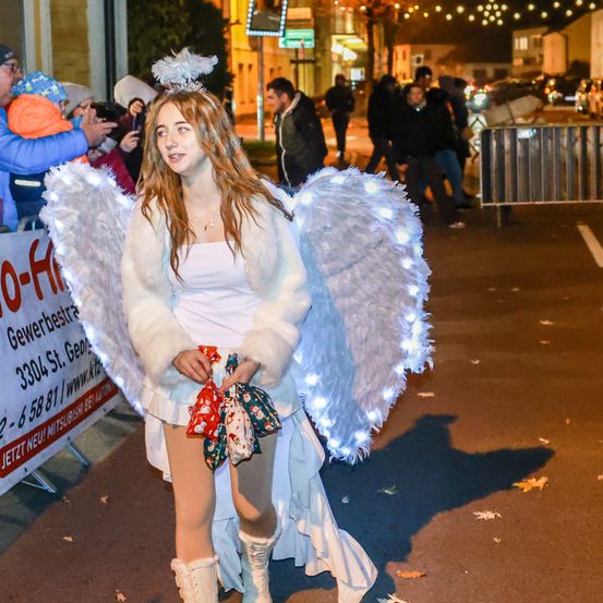 Eine junge Frau, als Engel verkleidet, geht in einer Parade, beleuchtet von beleuchteten Engelsflügeln, bei Nacht.