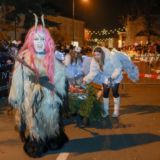 Eine Person in einem Monsterkostüm mit rosa Haaren und Hörnern schiebt einen Wagen mit einem Kranz, während eine Frau in Engelsflügeln daneben steht.