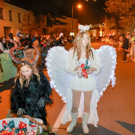 Zwei Kinder in Kostümen gehen bei Nacht in einer Straßenparade. Eines trägt ein Engelkostüm mit Flügeln und einem Heiligenschein, während das andere als Teufel verkleidet ist. Eine Menge schaut von den Seiten zu.