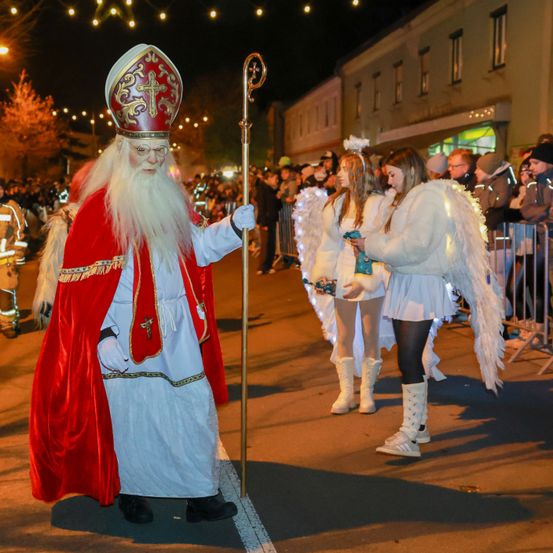Ein Mann, verkleidet als St. Nikolaus, führt eine Prozession auf einem Stadtplatz an, umgeben von jungen Frauen in Engelskostümen. Zuschauer säumen die Straße und beobachten.
